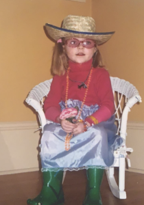 A small child wearing green galoshes and a wicker hat sits in a chair. 
